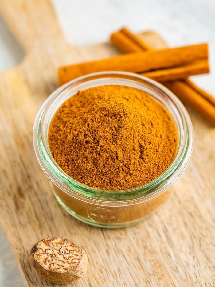 Small glass jar with pumpkin pie spice on a wood board and next to two sticks of cinnamon.