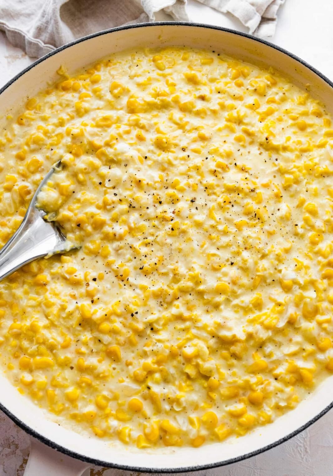 Creamed corn served in a white skillet with a silver spoon topped with freshly cracked pepper.