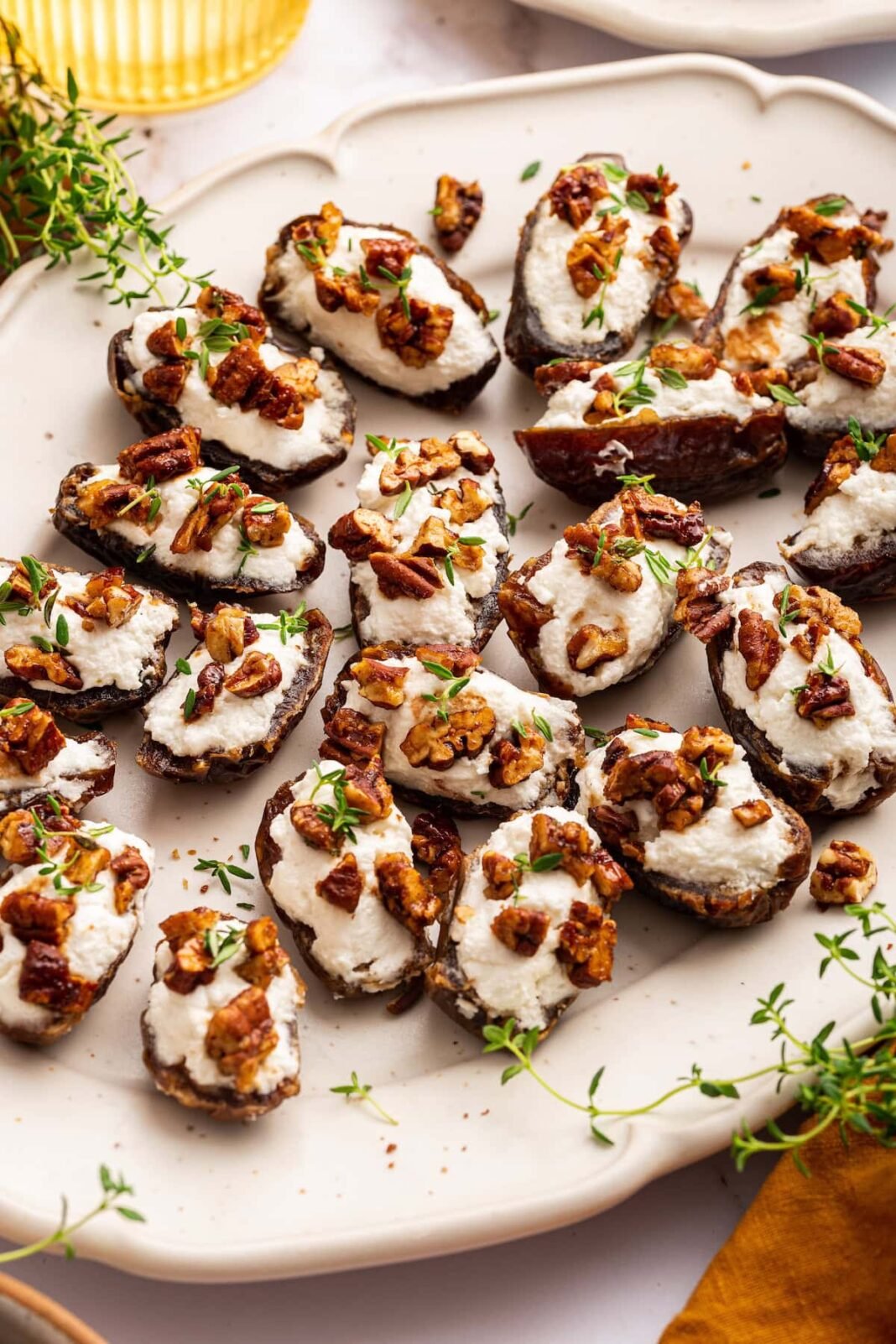 A large plate of multiple dates that are stuffed with goat cheese and topped with chopped pecans and fresh thyme.