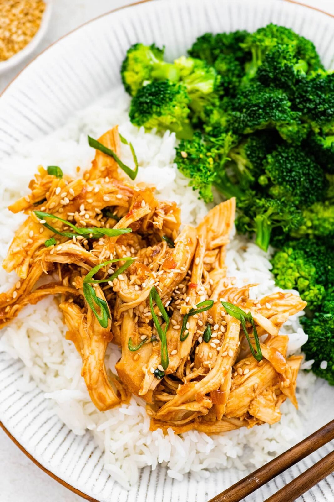 Slow cooker teriyaki chicken served on a plate with white rice and broccoli. The chicken is topped with sesame seeds and green onions.