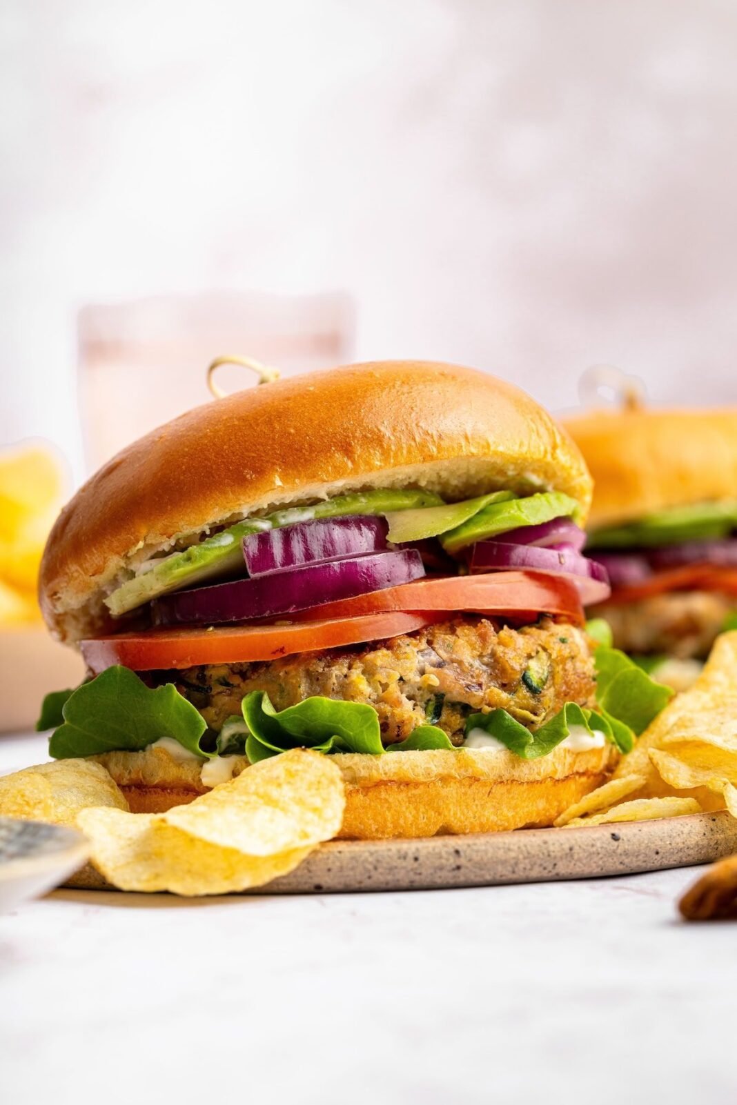A chicken burger served on a brioche bun topped with lettuce, tomato, red onion and avocado. The burger is served with a side of potato chips.